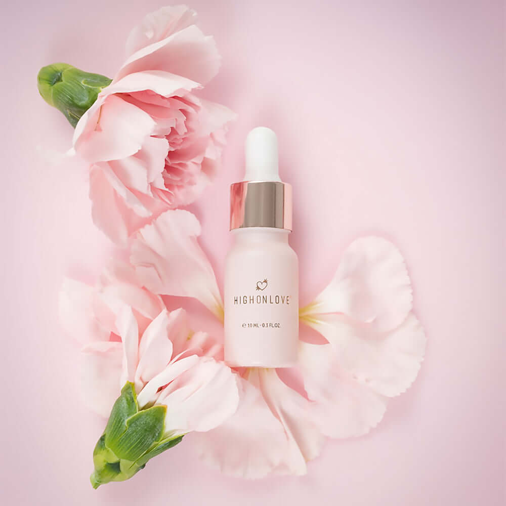 HighOnLove Mini Sensual Stimulating Oil bottle surrounded by soft pink flowers on a pastel background.