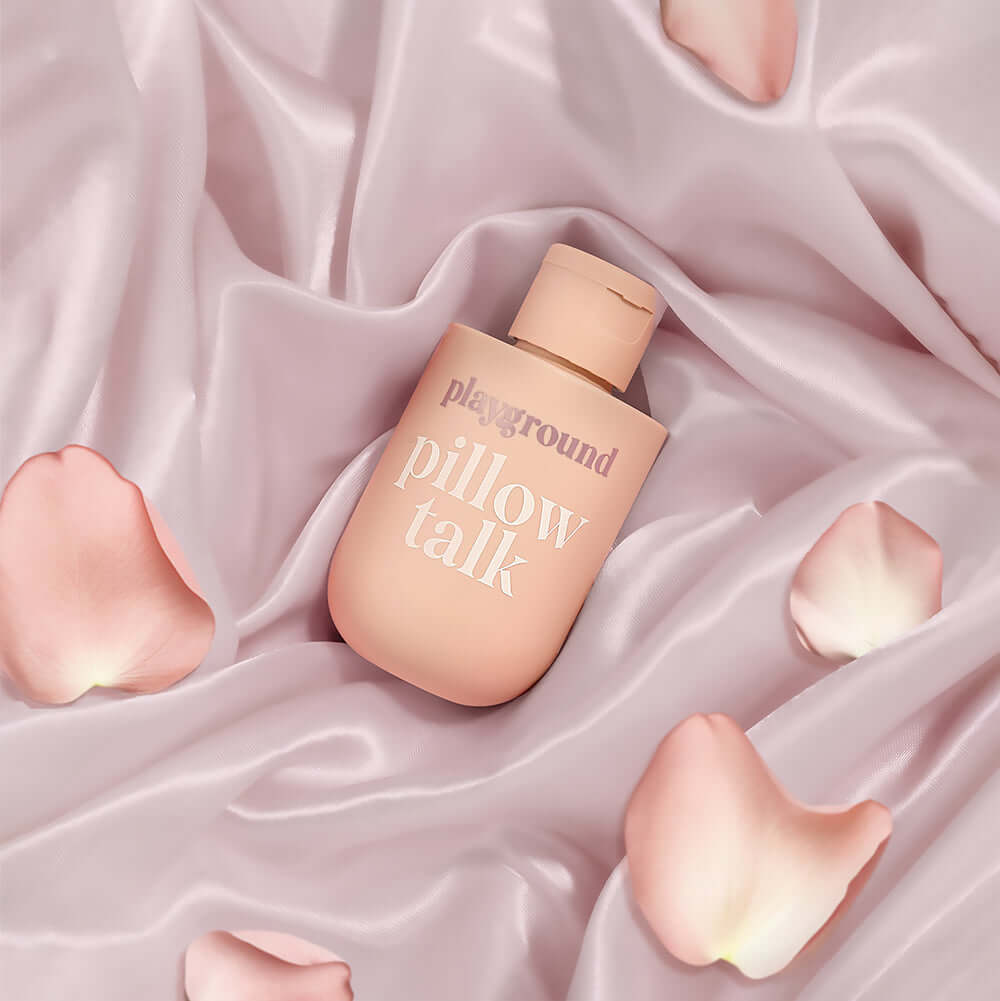 Playground Pillow Talk Water-Based Personal Lubricant bottle on pink satin fabric with rose petals.