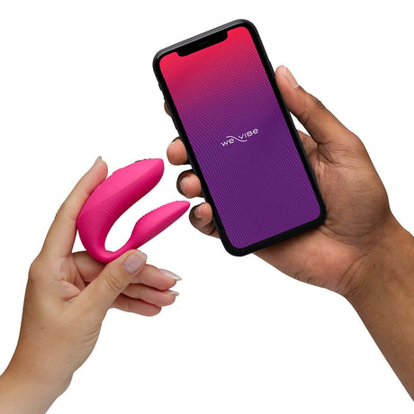 We-Vibe Chorus Pro Couples Massager Electric Pink held in one hand with smartphone displaying app in the other hand.