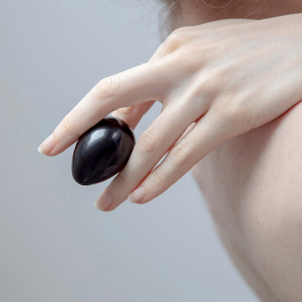 Chakrubs Xaga Yoni Egg Obsidian non-drilled held delicately between fingers, showcasing its smooth surface and design.