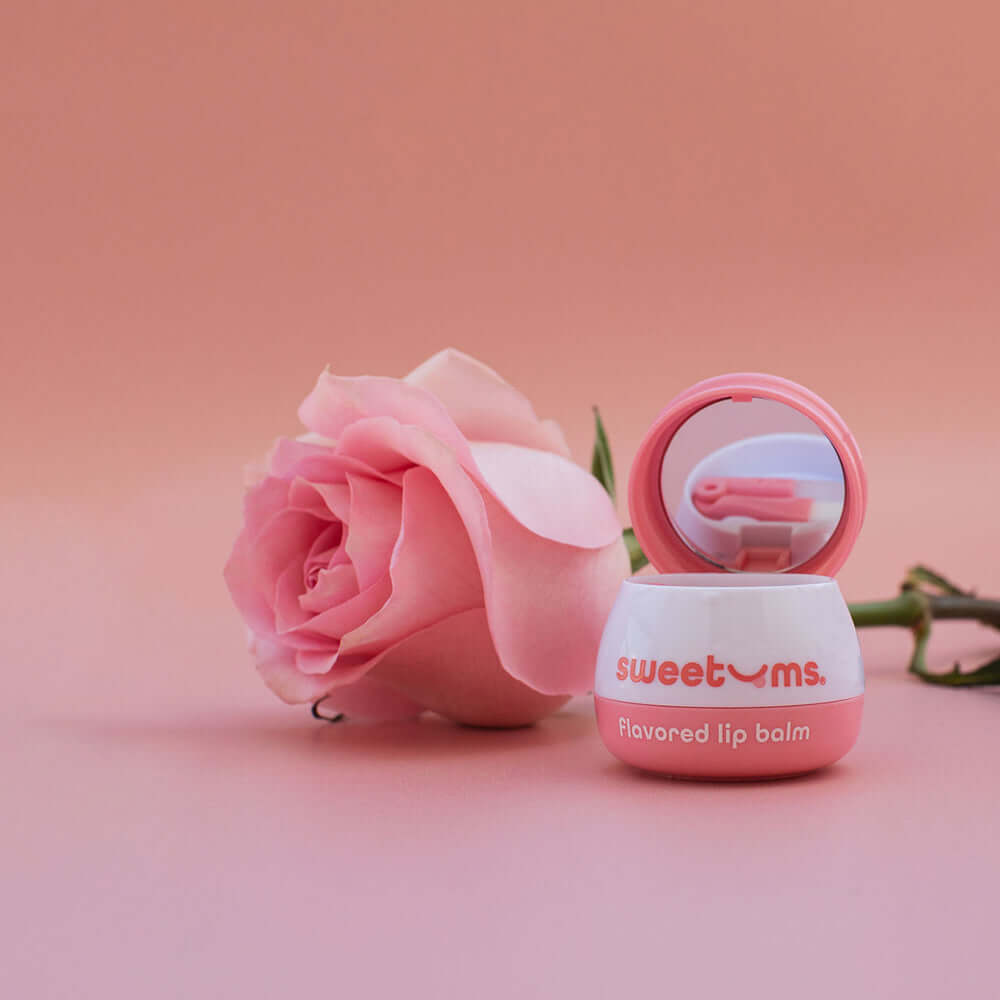 Sweetums Flavored Lip Balm Sweet Rose with a pink rose on a soft pink background, showcasing its moisturizing properties.