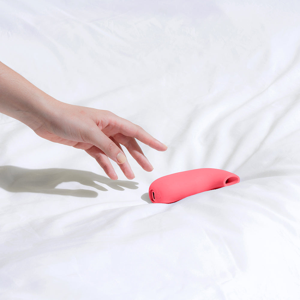 Dame Aer Suction Massager Coral resting on a white bedspread with a hand reaching for it.