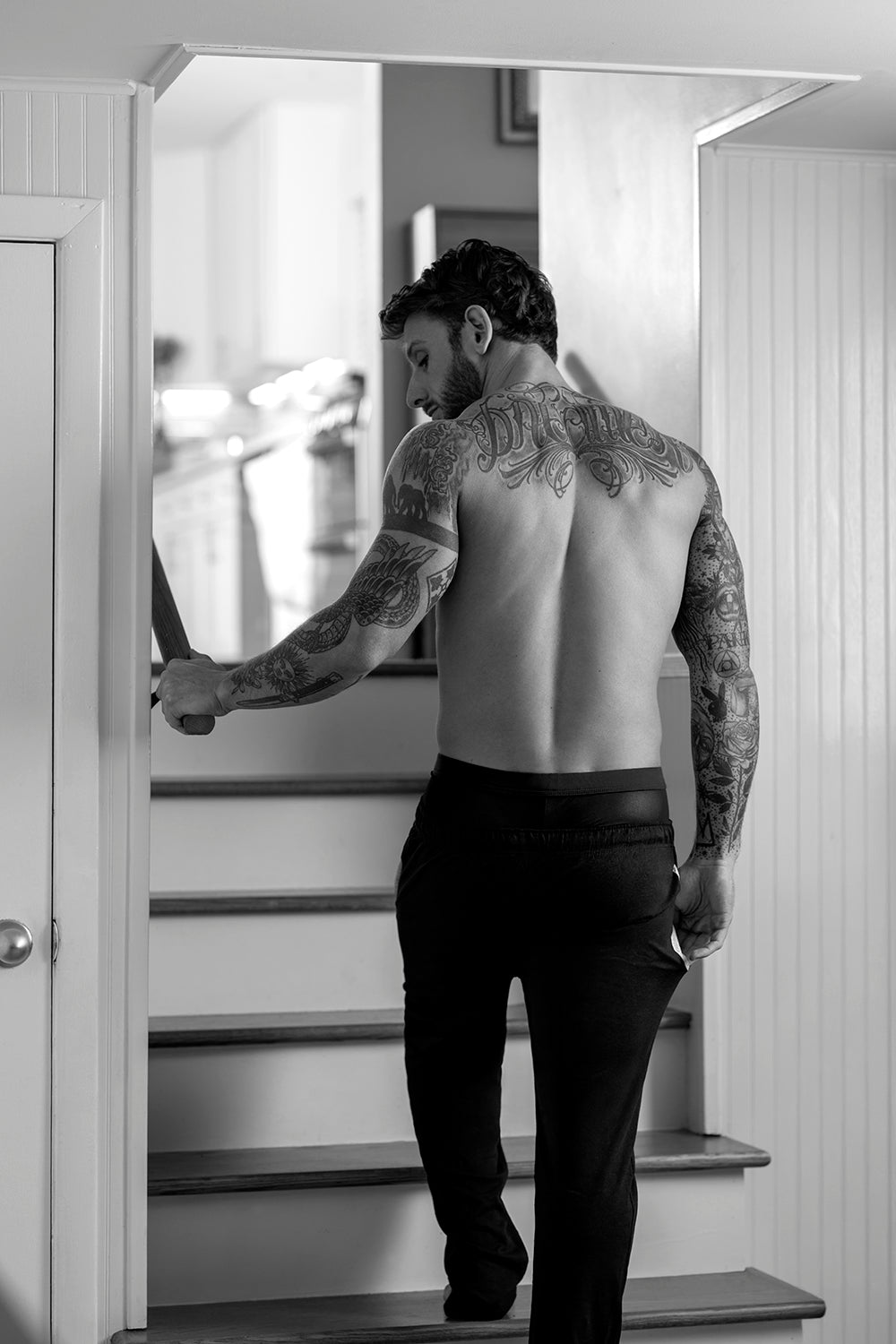 Man with tattoos wearing black pants walking up stairs in a modern home.