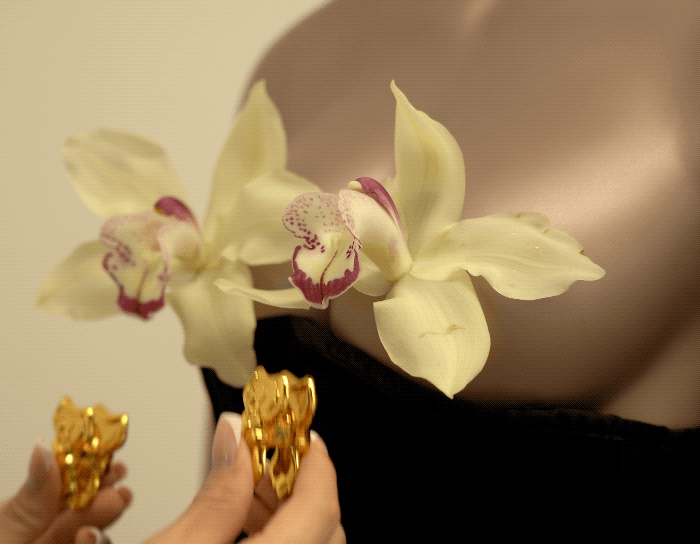 Hands holding golden bowknot nipple clamps with orchid flowers on a mannequin, showcasing elegant design and interaction.