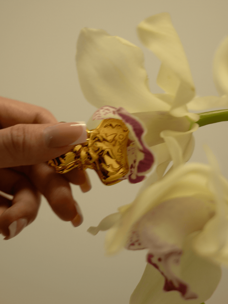 Hand holding a gold bowknot binder clip style nipple clamp near a white orchid flower.