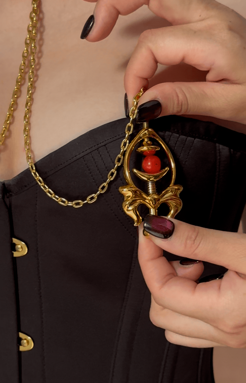 Close-up of a hand holding gold Pulling Chain Nipple Clamps with red accent, showcasing intricate design and chain details.