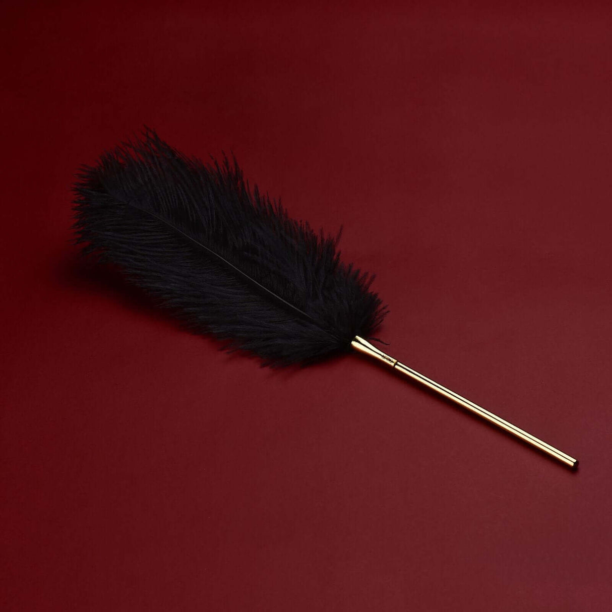 Soft black feather tickler with gold handle on a red background for sensual play and heightened arousal.