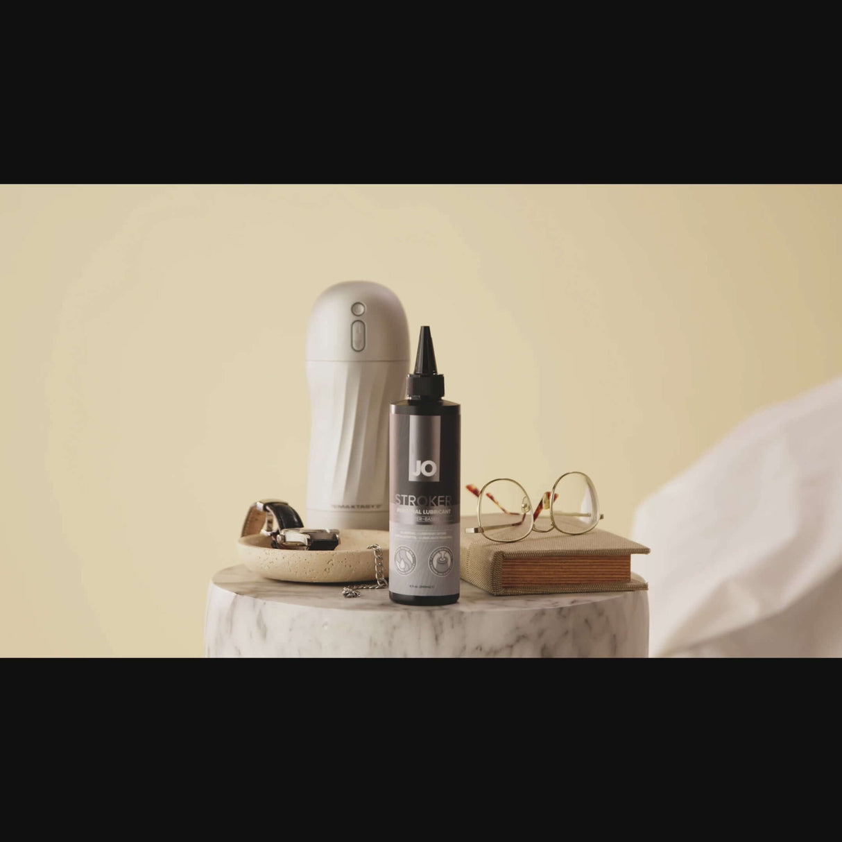 JO Stroker Lubricant 4 Oz displayed next to a stroker, rings, and glasses on a marble table.