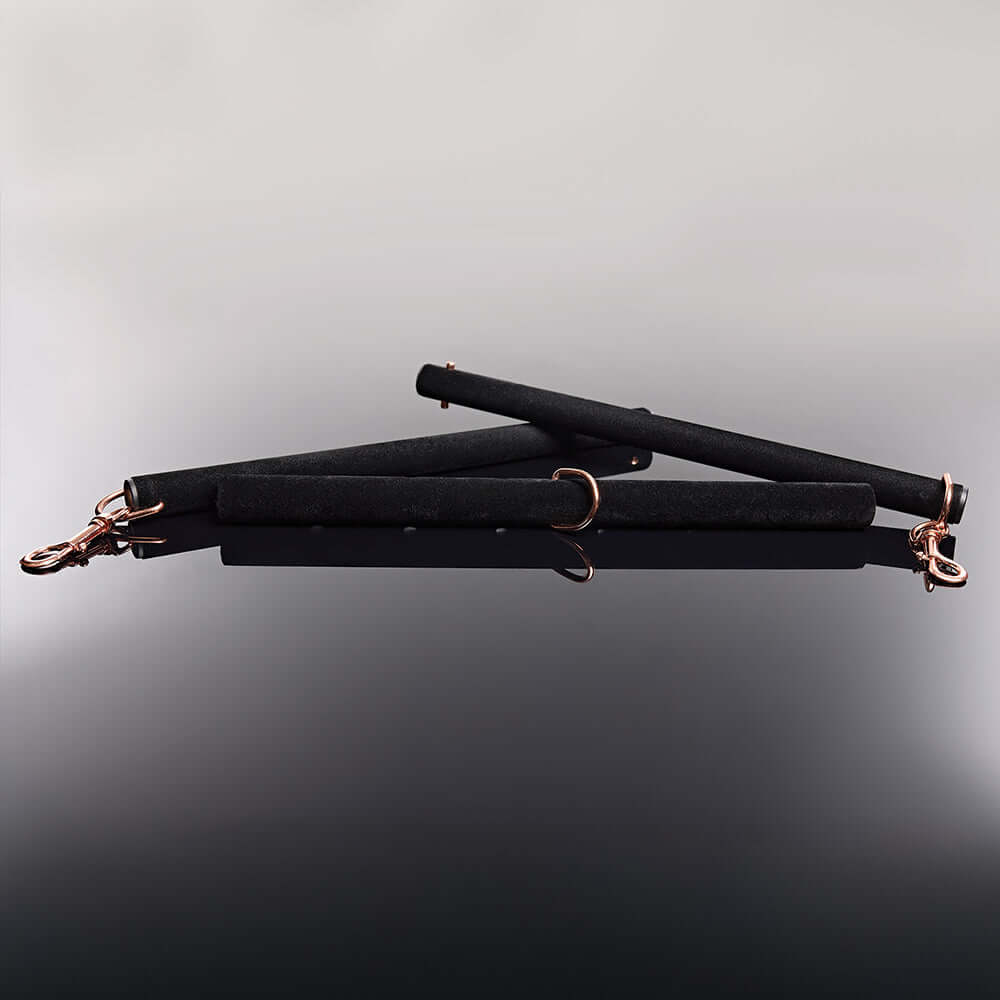Coquette Pleasure Spreader Bar Black with velvet finish and metal accents placed on a reflective surface.