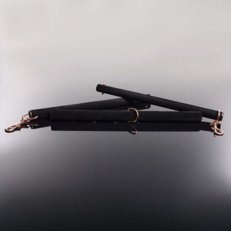 Coquette Pleasure Spreader Bar Black with velvet finish and metal accents placed on a reflective surface.