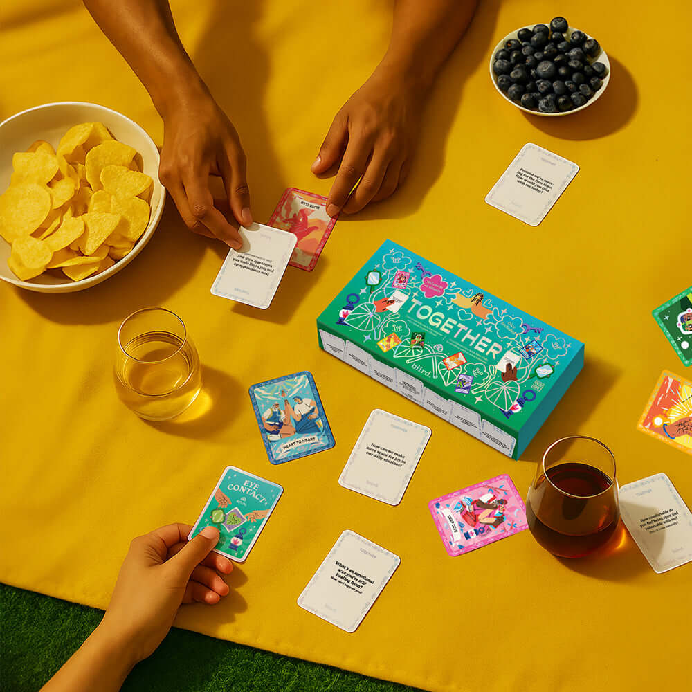 Biird x Jouissance Club Together Couples Card Game set on a yellow table with snacks and conversation cards.