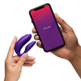 Person holding the We-Vibe Chorus Pro Cosmic Purple and a smartphone with the We-Vibe app.
