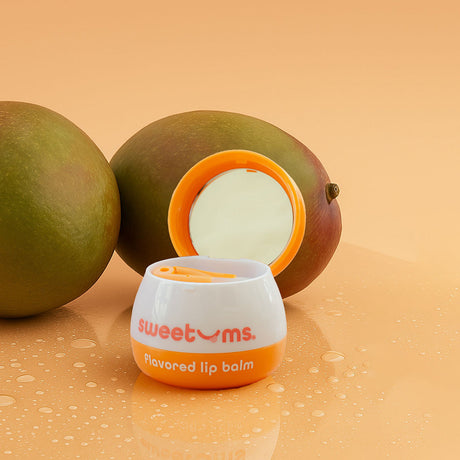Sweetums Flavored Lip Balm Mango compact next to fresh mangoes on a reflective surface, highlighting the product's tropical appeal.