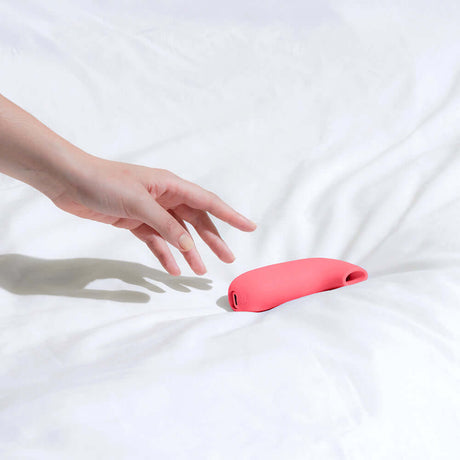 Dame Aer Suction Massager Coral resting on a white bedspread with a hand reaching for it.