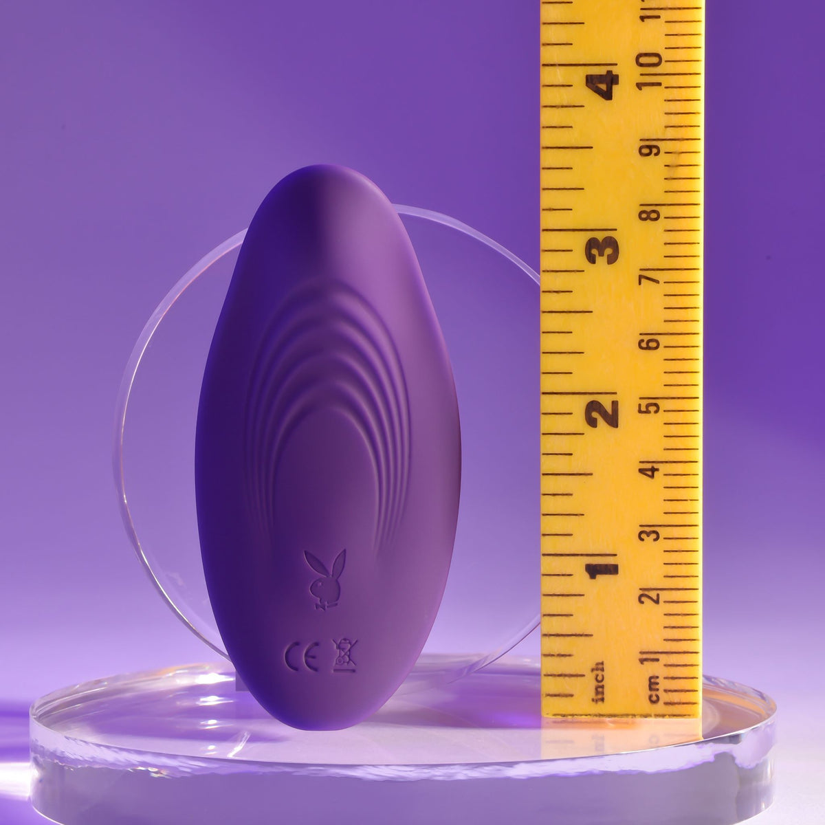 Playboy Our Little Secret Panty Vibrator Purple displayed next to a measuring tape on a clear surface.