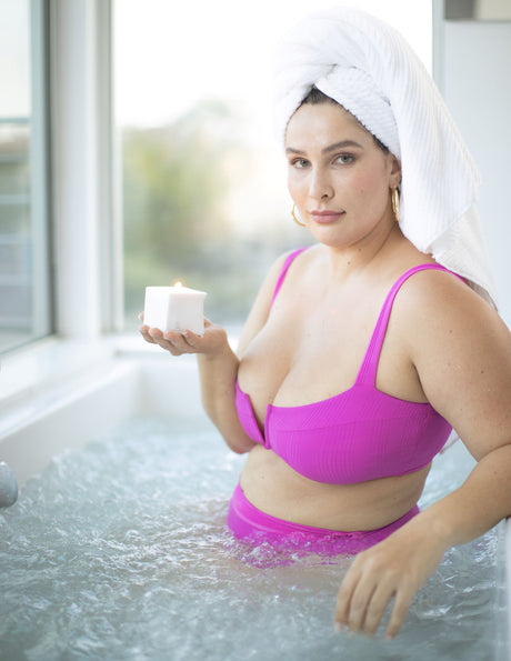 Woman holding a lit candle while relaxing in a bathtub, showcasing the soothing ambiance of Jimmyjane Natural Massage Oil Candle 4.5 Oz Santal.
