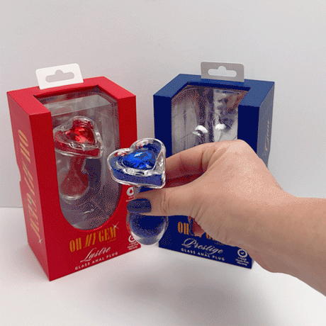 Person holding the Oh My Gem Prestige Glass Anal Plug Blue between red and blue packaging.