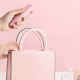 Hand holding pink Sweet Secret Massager above a pink handbag with a soft pink background.