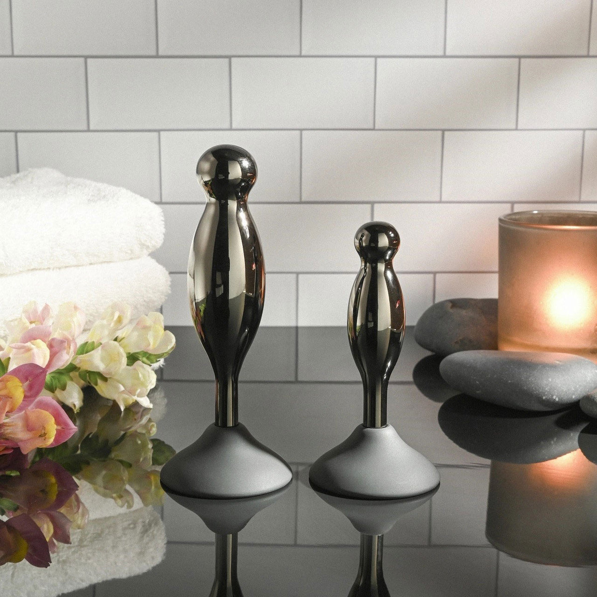 Evolved Joy Sticks 2 Piece Anal Plug Set Black Chrome displayed with towels, flowers, and candles.