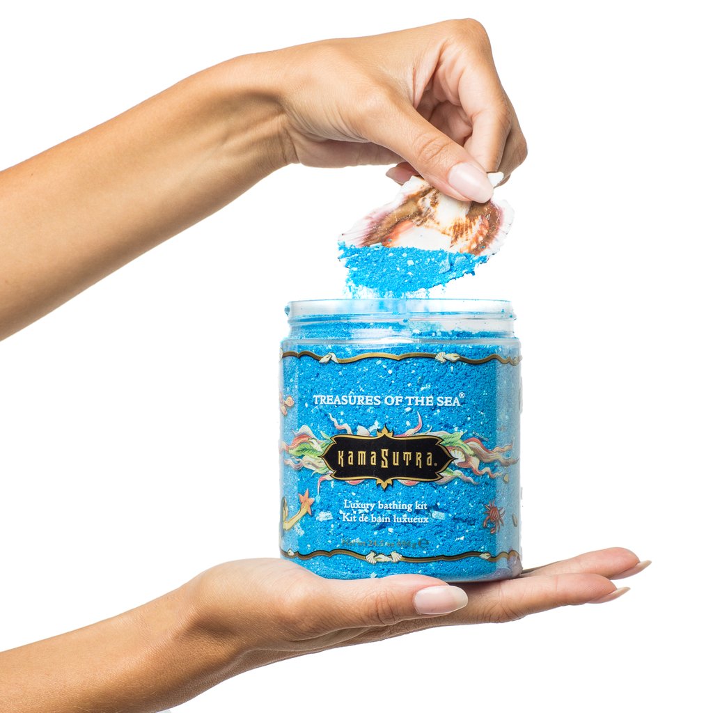 A person pouring blue crystal bath salt from a jar labeled Kama Sutra Treasures Of The Sea Bath Salt.